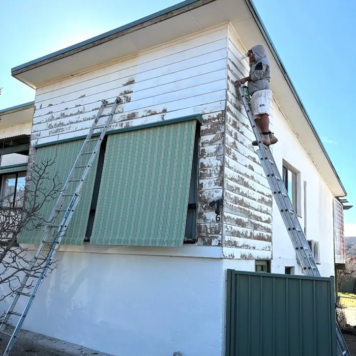 Peeling, patchy weatherboard on an upstairs corner, with a ladder against the wall and a green striped awning.
