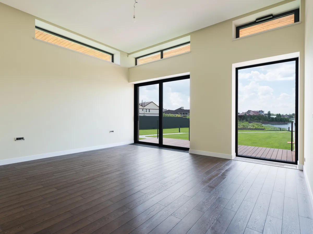 Empty new-build living room with pale walls, white trim and dark timber floor, opening onto a garden through full-height glass doors.